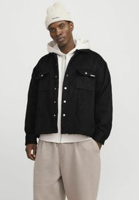 Veste en polaire noire avec deux poches avant, boutons-pression et design court, superposée sur un sweat à capuche gris clair et un pantalon crème.