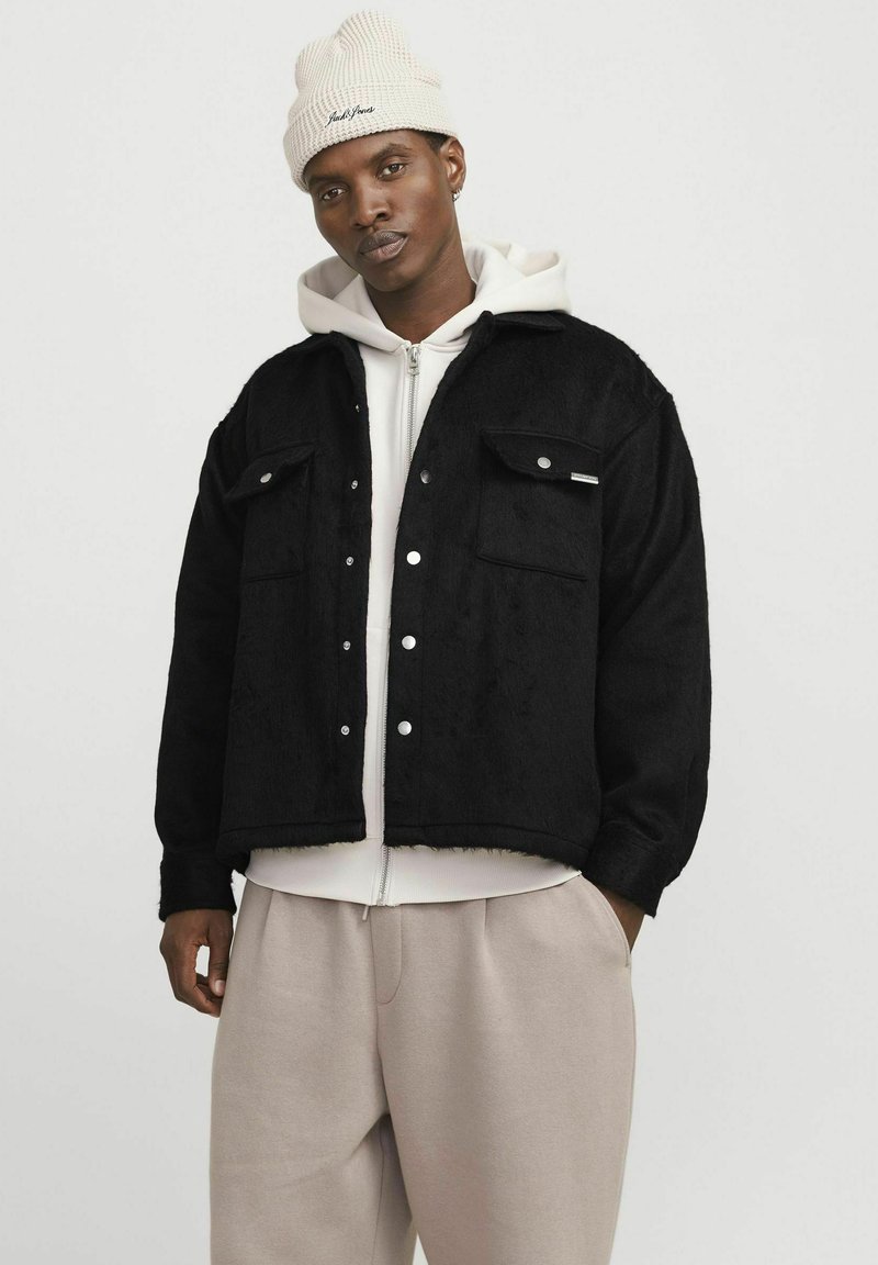 Veste en polaire noire avec deux poches avant, boutons-pression et design court, superposée sur un sweat à capuche gris clair et un pantalon crème.