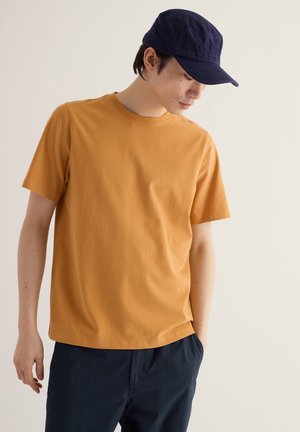 Jeune homme portant une chemise jaune moutarde à manches courtes, un pantalon bleu marine et une casquette bleue marine, regardant vers le bas avec une main dans la poche, sur un fond uni.