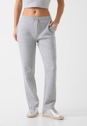 Kvinde iført lysegrå joggingbukser og metallic sølv sneakers, stående med den ene hånd i lommen mod en ensfarvet hvid baggrund.