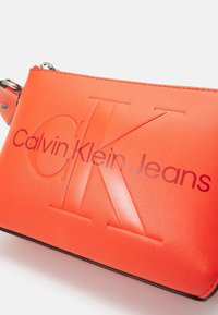 Wristlet Calvin Klein Jeans in un vivace arancione, realizzato in materiale sintetico liscio, con logo in rilievo e chiusura zip.