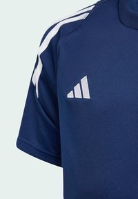 Närbild av en marinblå sporttröja med vita ränder på axeln och en liten broderad logotyp på bröstet.