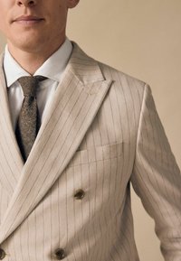 Double-breasted beige suit jacket with light purple pinstripes, notched lapels, and a chest pocket. Textured brown tie complements the look.