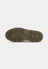 Semelle d'une botte en caoutchouc marron avec des crampons profonds, empreinte de logo et petit détail hexagonal bleu près du talon.