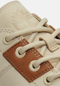 Primer plano de cordones beige de botas pasados por ojales redondos y un parche de cuero marrón con costuras visibles y un logo de árbol en relieve.