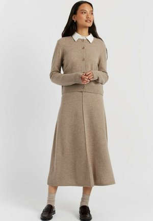 Vrouw draagt een beige gebreid vest met knopen, bijpassende midi rok, beige sokken, zwarte loafers en een wit overhemd met kraag, staand tegen een effen achtergrond.