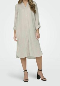 Donna che indossa un vestito camicia midi beige con bottoni, maniche arrotolate e sandali con tacco a blocco neri e cinturino alla caviglia.