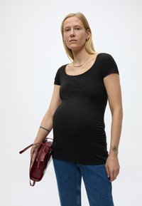 Hosszú szőke hajú kismama fekete felsőt és kék farmert visel, egy sötétvörös táskát tart egyszerű fehér háttér előtt.