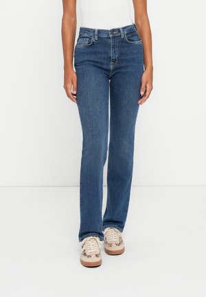 Dark blue denim jeans with a fitted design, straight leg, and five-pocket styling, paired with light-colored sneakers.