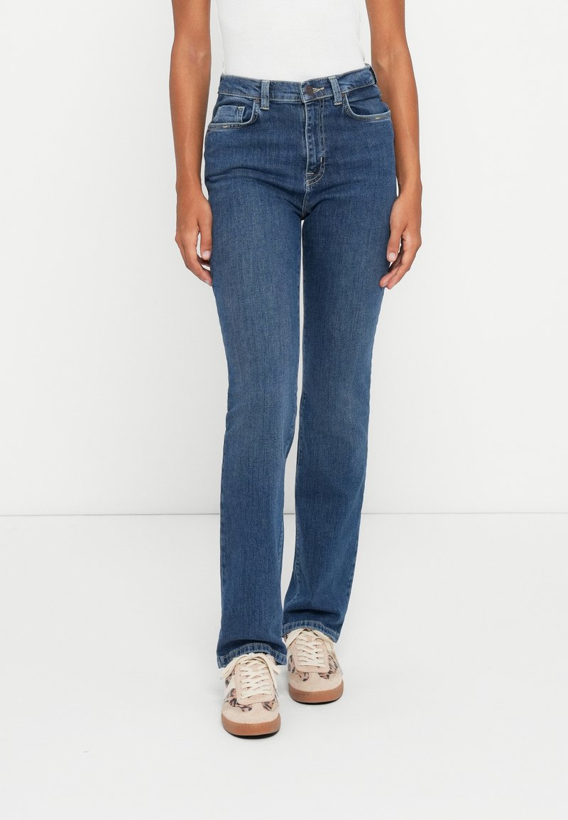 Jeans de mezclilla azul oscuro con un diseño ajustado, pierna recta y estilo de cinco bolsillos, combinados con zapatillas de color claro.
