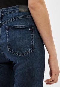 Tuvplāns uz personas apakšējo muguras daļu un roku, valkājot tumši zilas džinsa bikses ar šūtu aizmugurējo kabatu un melnu ādas zīmolu uzlīmi.