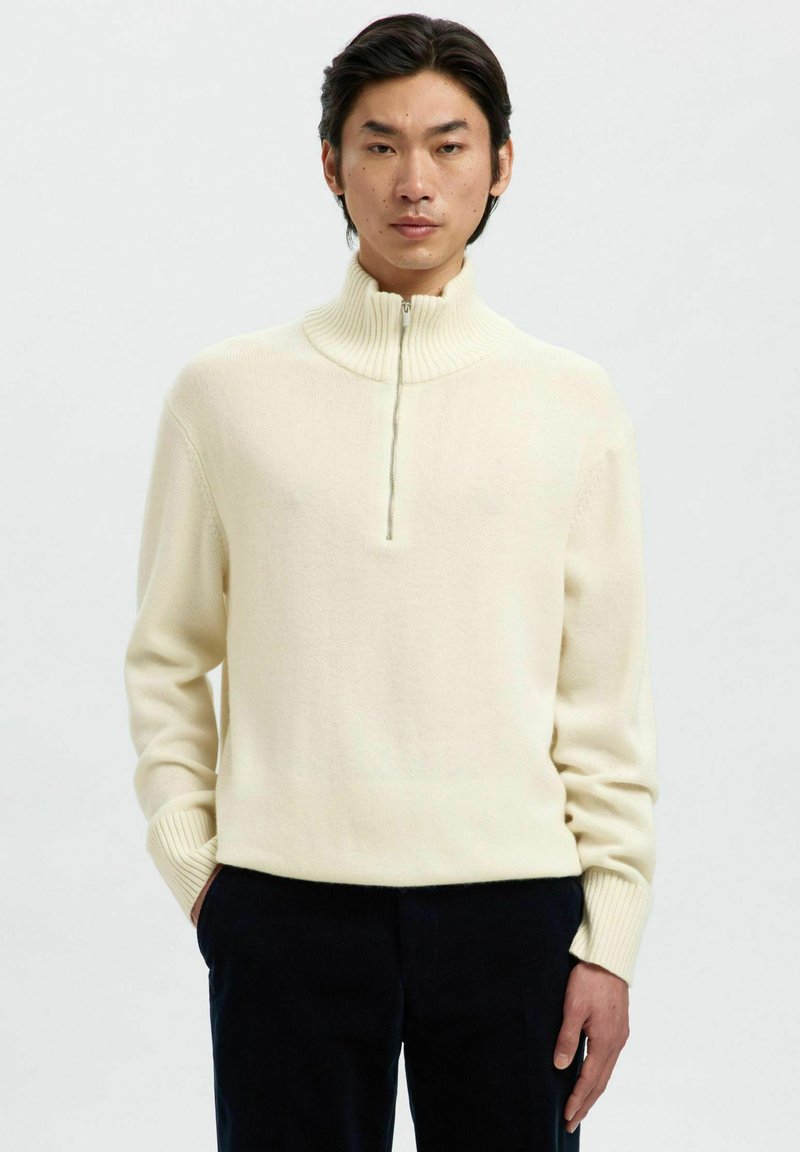 Cremfarbener Strickpullover mit hohem Kragen, einem Viertel-Reißverschluss vorne und gerippten Bündchen, hergestellt aus weichem, strukturiertem Stoff.