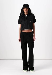 Camisa preta cropped com mangas curtas e bolsos no peito, emparelhada com calças pretas de perna larga e um chapéu tipo balde preto correspondente. Sapatos com sola branca.