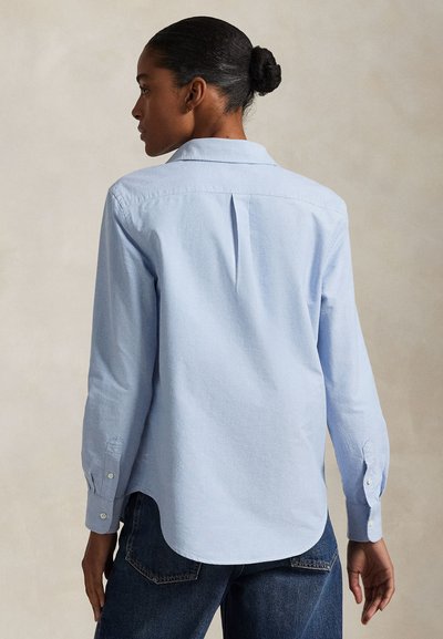 Woman with dark hair in a bun wearing a light blue long-sleeve shirt and dark jeans, standing and facing away from the camera.