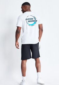 Camiseta de algodón blanca con gráfico circular "ELEVEN DEGREES" en azul en la parte posterior, combinada con pantalones cortos negros. El modelo lleva zapatillas blancas.