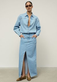 Šviesiai mėlyna džinsinė apranga, sudaranti iš per didelio mygtukų marškinių ir ilgos sijono su priekinėmis plyšiais, derinama su rudais zomšiniais batais.