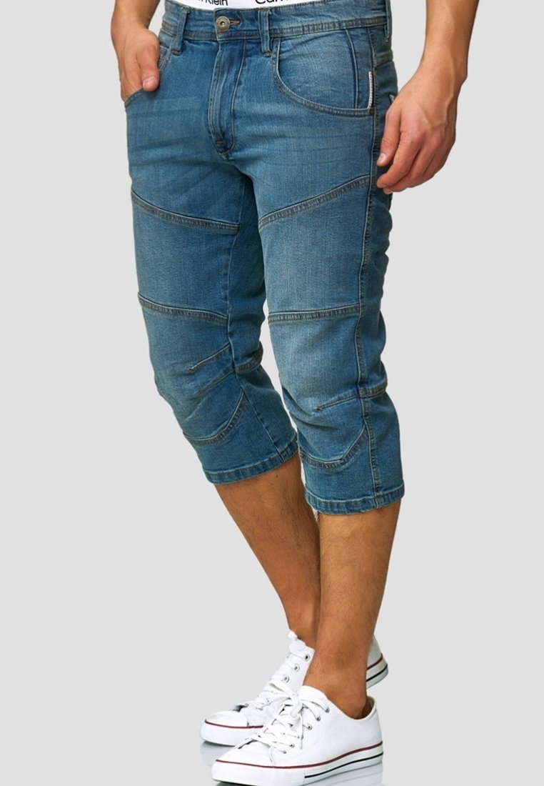 Homme portant un pantalon en jean bleu raccourci avec des détails cousus et des baskets blanches, debout avec une main dans la poche.