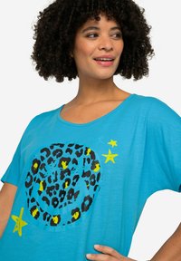 Camiseta turquesa de corte holgado, con un gráfico de estampado de leopardo negro y estrellas amarillas. Tejido suave con cuello redondo.