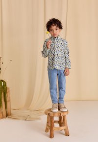 Kleurijk langhemd met een bladerenpatroon, gecombineerd met lichtblauwe jeans en beige schoenen, staand op een houten kruk.