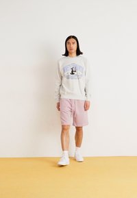 Felpa grigia chiara con grafica "Detroit", abbinata a pantaloni corti rosa chiaro e sneakers bianche. Sfondo semplice con pavimento giallo.