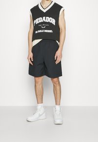 Černý bezrukávový svetr s nápisem "PEGADOR", béžovými akcenty; v kombinaci s černými šortkami a bílými vysokými teniskami.