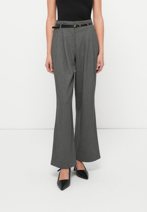 Nainen pukeutunut korkeavyötäröisiin harmaisiin leveälahkeisiin housuihin, joissa on laskokset, musta vyö, musta hihaton paita ja mustat teräväkärkiset korkokengät, seisoo valkoisen seinän vierellä.