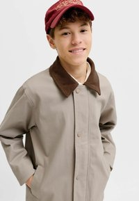 Adolescent portant une veste beige avec col marron et une casquette rouge, mains dans les poches, souriant doucement devant un fond clair uni.