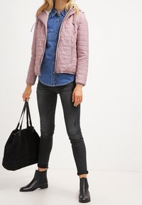 Světle růžová prošívaná bunda s kapucí, nošená přes denimovou košili. Černé úzké džíny a kotníkové boty. Černá kabelka dokresluje outfit.