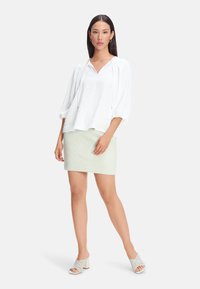 Weißer Pullover mit V-Ausschnitt und Kordelzug, kombiniert mit einem strukturierten, hellgrünen Minirock und offenen, weißen Blockabsatzsandalen.