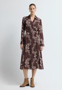 Lange bruine jurk met een wikkelontwerp, een ingewikkeld gebroken wit paisley-patroon, een kraag en een knoop ter hoogte van de taille. Paar lichtbruine enkellaarsjes.