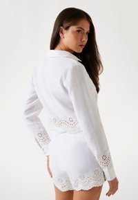 White linen crop jacket with a scalloped hem and floral eyelet detailing; paired with matching white shorts, emphasizing texture and design.