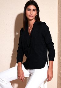 Black long-sleeve button-up top with two chest pockets, textured fabric, and a relaxed fit, paired with white trousers.