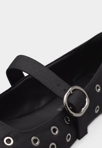 Μαύρο παπούτσι Mary Jane με υφασμάτινο λουράκι και μεταλλική αγκράφα, διακοσμημένο με ασημένια κρικάκια κατά μήκος της πλευράς.