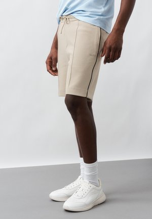 Άτομο που φοράει μπεζ σορτς με κορδόνι και μαύρη ρίγα, ανοιχτό μπλε πουκάμισο, λευκές κάλτσες και λευκά αθλητικά παπούτσια, στέκεται σε γκρι δάπεδο.