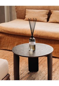 Tavolino da caffè rotondo nero con superficie texturizzata. Sopra vi è un diffusore in vetro con bastoncini neri, accanto a un divano beige e un tappeto intrecciato.