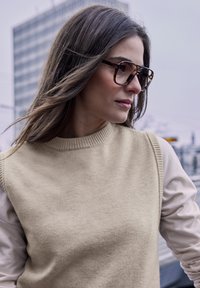 Pull en maille beige avec un col côtelé, associé à une chemise légère à manches longues. De grandes lunettes écaille de tortue ornent le visage.