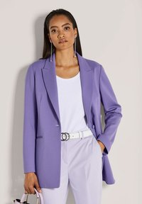 Vrouw in een lavendel blazer en broek met een witte top en riem, die een lavendel handtas vasthoudt, staande tegen een neutrale muur.