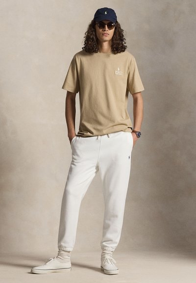 Άνδρας με μακριά σγουρά μαλλιά που φοράει μπέζ μπλουζάκι Polo Ralph Lauren, λευκές φόρμες, ναυτικό καπέλο, γυαλιά ηλίου και λευκά αθλητικά παπούτσια, στέκεται με τα χέρια στις τσέπες.