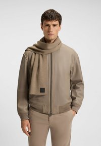 Beige bomberjakke med lynlås foran og ribbede manchetter, parret med et matchende tørklæde. Stoffet ser glat ud med en subtil tekstur.