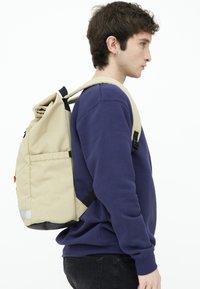 Rulltop ryggsäck i beige tyg, med justerbara svarta remmar, sidoficka och reflekterande detaljer vid basen.