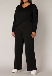 Zwarte langslavige top met V-hals, gecombineerd met wijde zwarte broeken. Model draagt witte sneakers. Zachte stof met een gladde textuur.