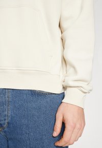 Krämfärgad hoodie i mjukt tyg med kängurufickor och ribbade ärmavslut. Bärs med blå denimjeans.