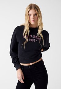 Czarna krótka bluza z nadrukiem "Guess Jeans 1981" w kolorze różowym. Charakteryzuje się szerokimi rękawami i miękką teksturą. Połączona z czarnymi, wysokim stanem jeansami.