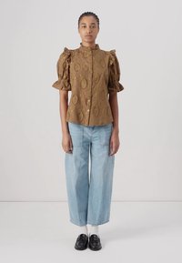 Blusa marrone con maniche a palloncino e motivi foglia texture, chiusura con bottoni, abbinata a jeans a gamba larga blu chiaro e scarpe nere.