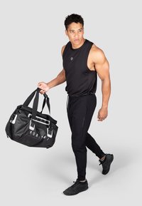 Schwarze ärmellose Sporttop und tapered Jogginghosen, getragen von einem Mann, der eine schwarze Sporttasche mit weißen Details hält, vor einem grauen Hintergrund.