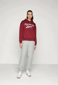 Sweatshirt maroon da Reebok com um logo branco, combinado com calças de moletom cinza claro. O sweatshirt apresenta um bolso frontal e cordões. Ténis brancos completam o visual.
