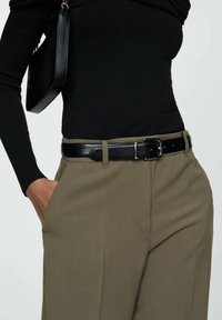 Persoon in een zwart langemouwen top, ingestopt in een hooggewaaid olijfgroene broek met een zwarte leren riem. Hand in de zak en de schouderband van een zwarte tas zichtbaar.