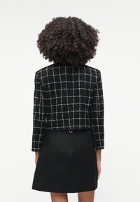 Sort cropped blazer med et gittermønster i hvid, som har lange ærmer og en tekstureret overflade, parret med en matchende sort nederdel.