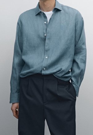Man wearing a blue button-up shirt over a white t-shirt and black pants with one hand in his pocket against a plain background.