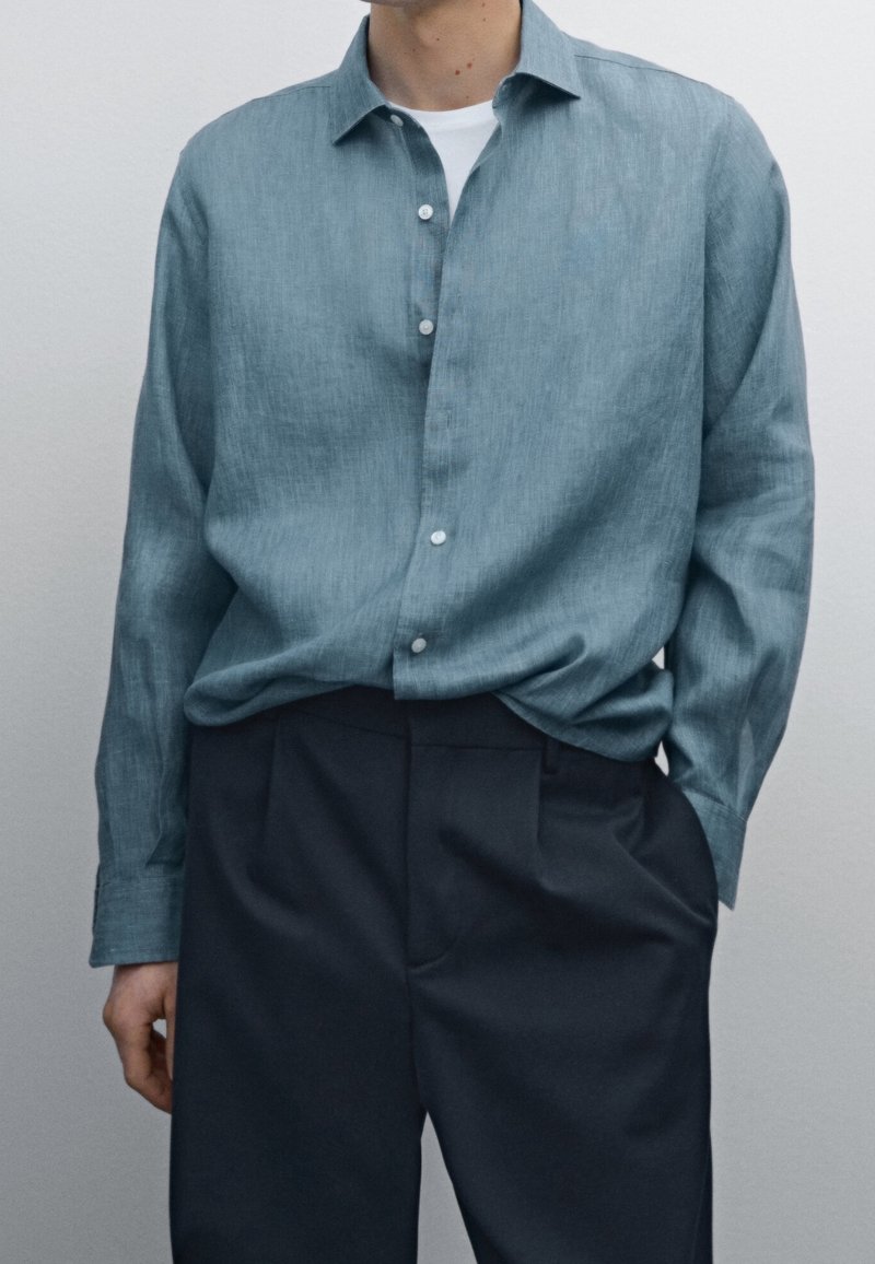Homme portant une chemise bleue boutonnée sur un t-shirt blanc et un pantalon noir, une main dans la poche, sur un fond uni.